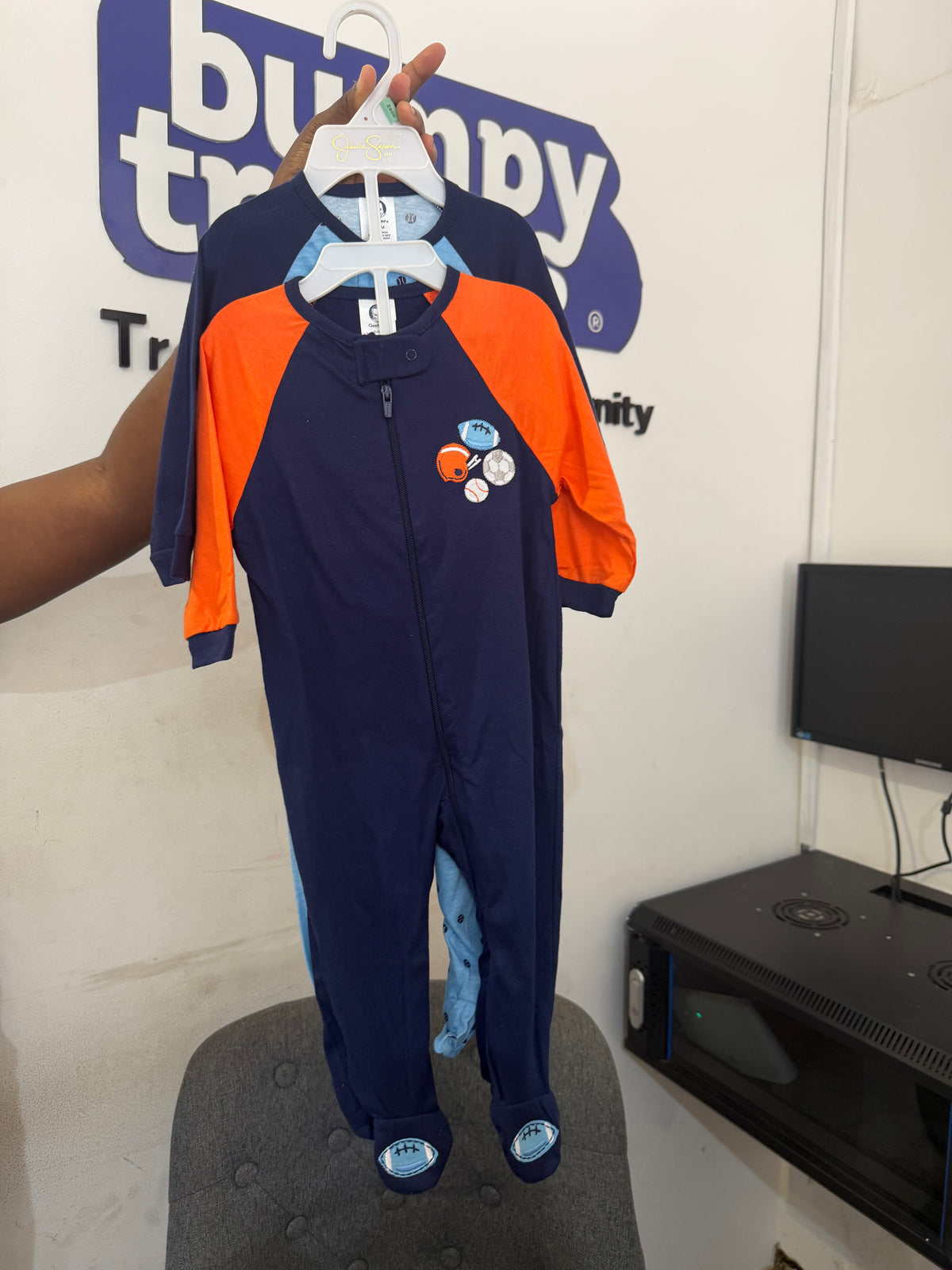 A baby one-piece footed sleeper in navy blue with bright orange sleeves, held up on a hanger by a hand. The sleeper features a front zipper, a small space-themed graphic on the chest, and blue foot covers.