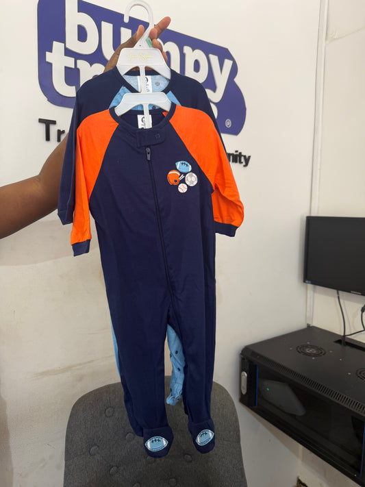 A baby one-piece footed sleeper in navy blue with bright orange sleeves, held up on a hanger by a hand. The sleeper features a front zipper, a small space-themed graphic on the chest, and blue foot covers.