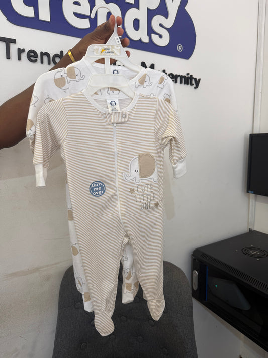 A baby footed sleeper in beige and white stripes with a cartoon elephant and the words 'WE LOVE LITTLE ONES' is held up on a hanger by a person's hand. A second matching sleeper is behind it.